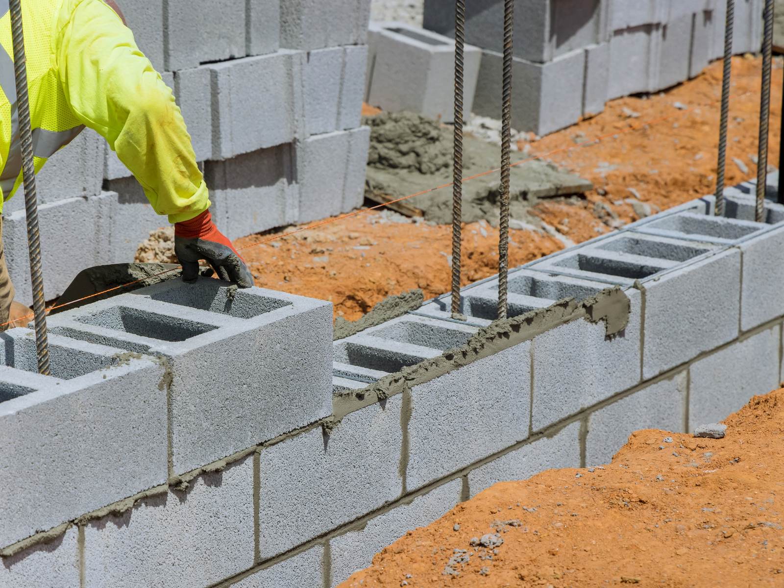Pedreiro de luvas assentando blocos de concreto com vergalhões verticais de espera em muro de arrimo com bloco de concreto